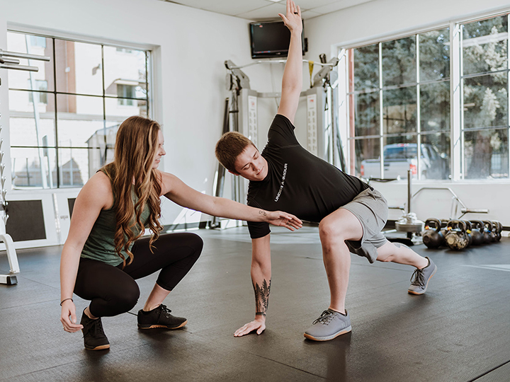 Female personal trainer instructing male client on a yoga pose inside a gym.